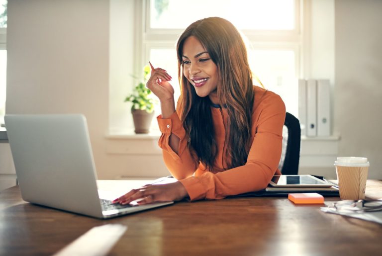 woman at desk working on laptop holding pen and smiling