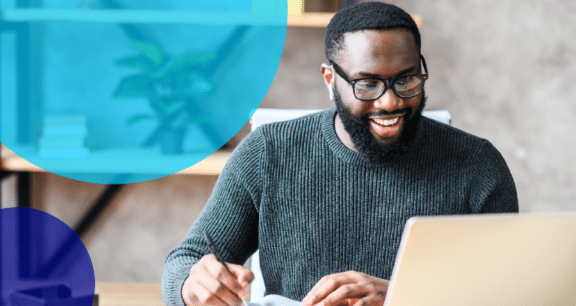 man writing on paper and looking at laptop smiling