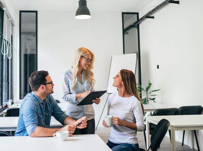3 people sitting at a desk holding coffee and pointing to a notebook