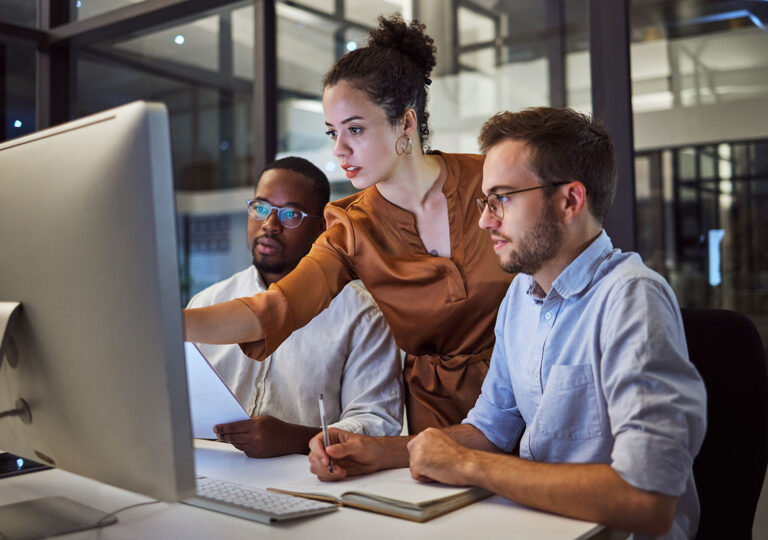 3 people sitting at desk looking and pointing at a desktop